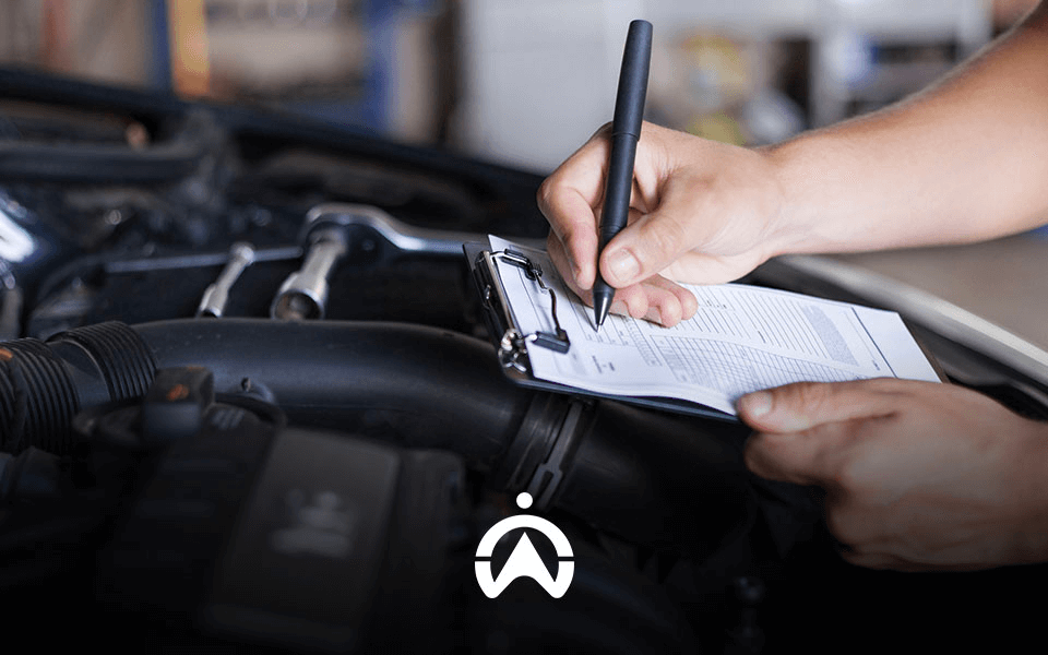 A mechanic notes observations on a clipboard while inspecting a car engine in a garage.