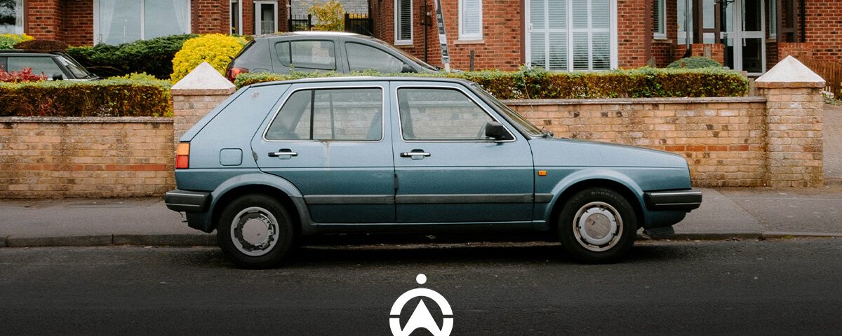 A vintage blue car parked on a residential street, with brick houses and hedges in the background.