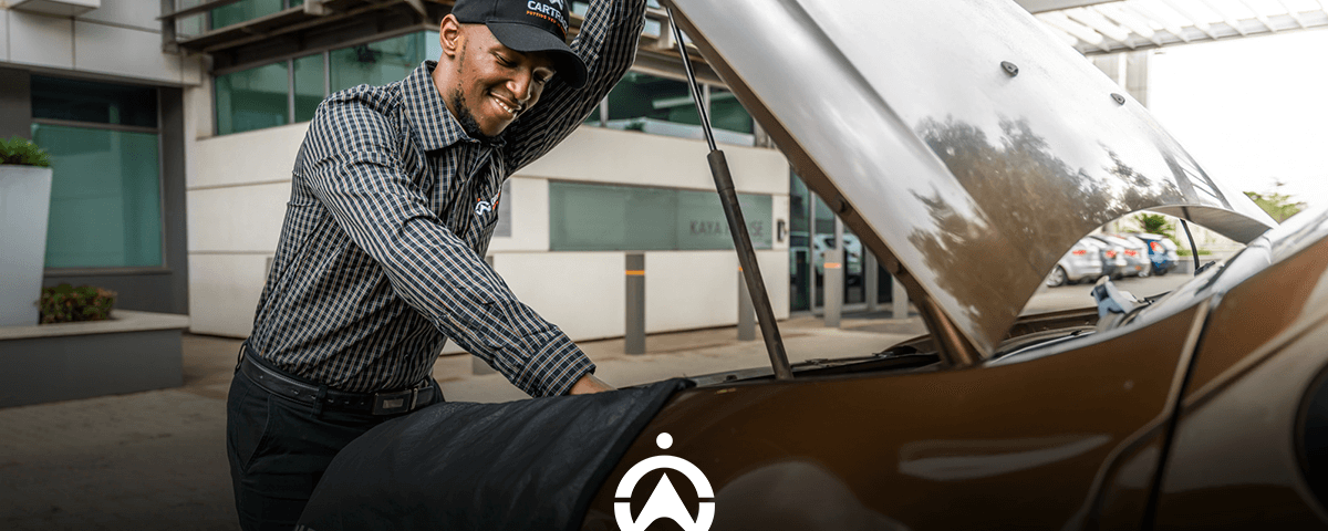 A technician inspects the engine of a brown car with the hood raised, parked outside a modern building.
