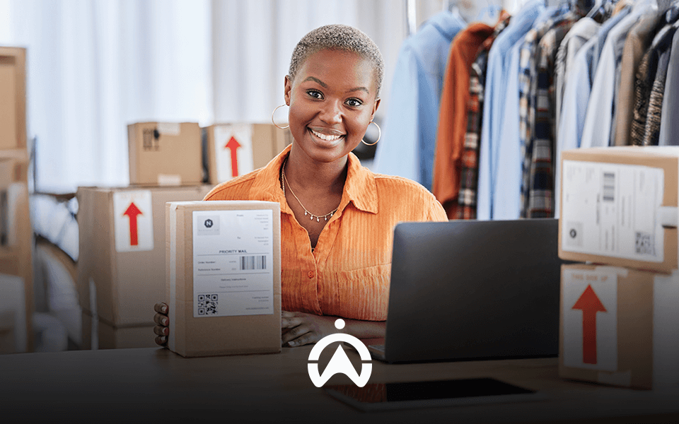 A person in an orange shirt holds a package in front of a laptop, surrounded by cardboard boxes and clothing on hangers.