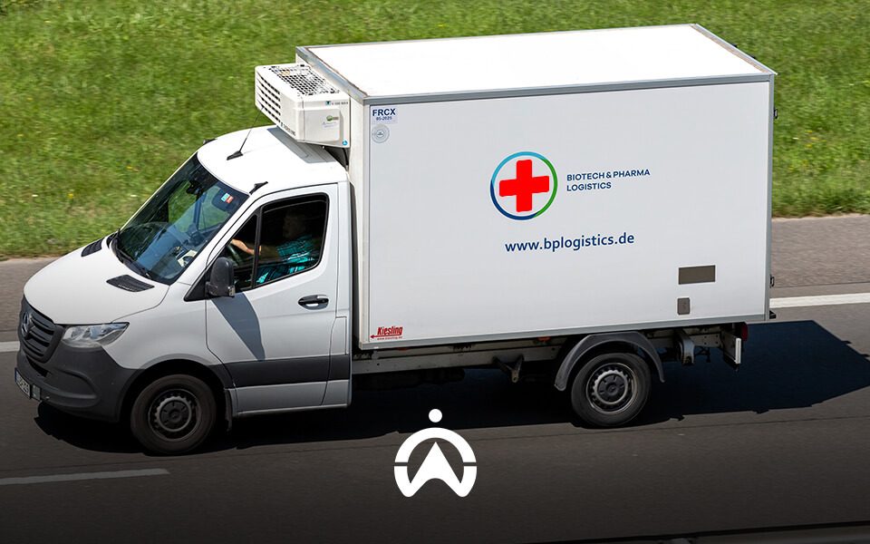 A white refrigerated logistics truck drives on a highway, displaying a red cross logo and "Biotech & Pharma Logistics" branding.