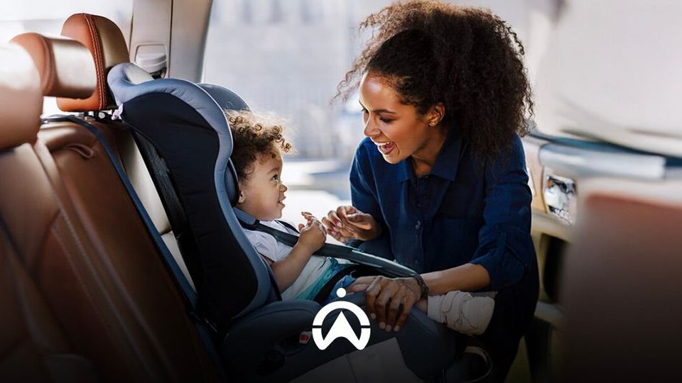 A woman interacts with a child securely seated in a car seat, surrounded by leather car interior, emphasizing safety and care.
