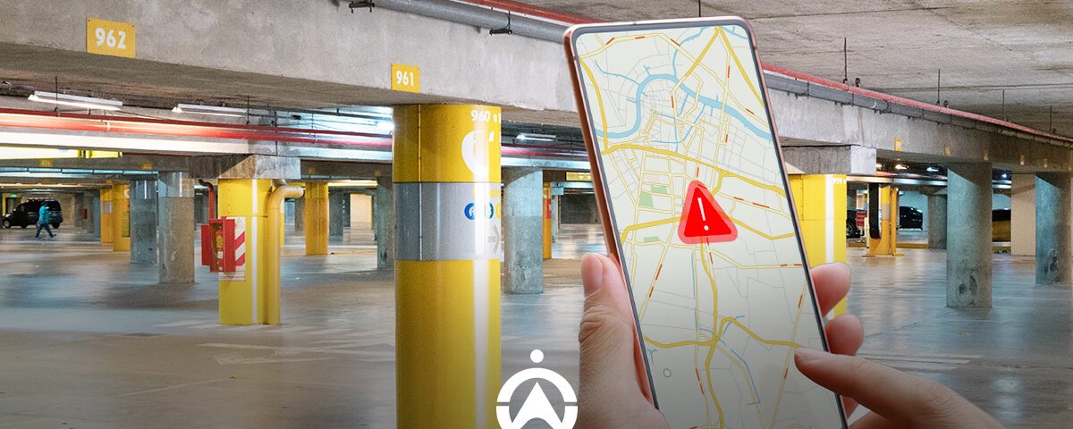 A person holds a smartphone displaying a map with a warning icon in a spacious, illuminated parking garage with yellow pillars.