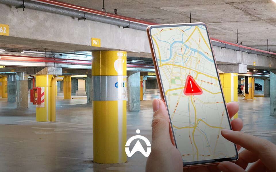 A person holds a smartphone displaying a map with a warning icon in a spacious, illuminated parking garage with yellow pillars.