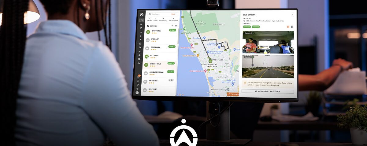 A woman views a detailed map and live camera feed on a monitor, monitoring routes and drivers for a logistics service.