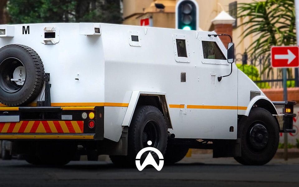 A white armored vehicle with yellow stripes parked on a street, displaying a distinctive wheel mounted on the rear. Traffic lights visible.