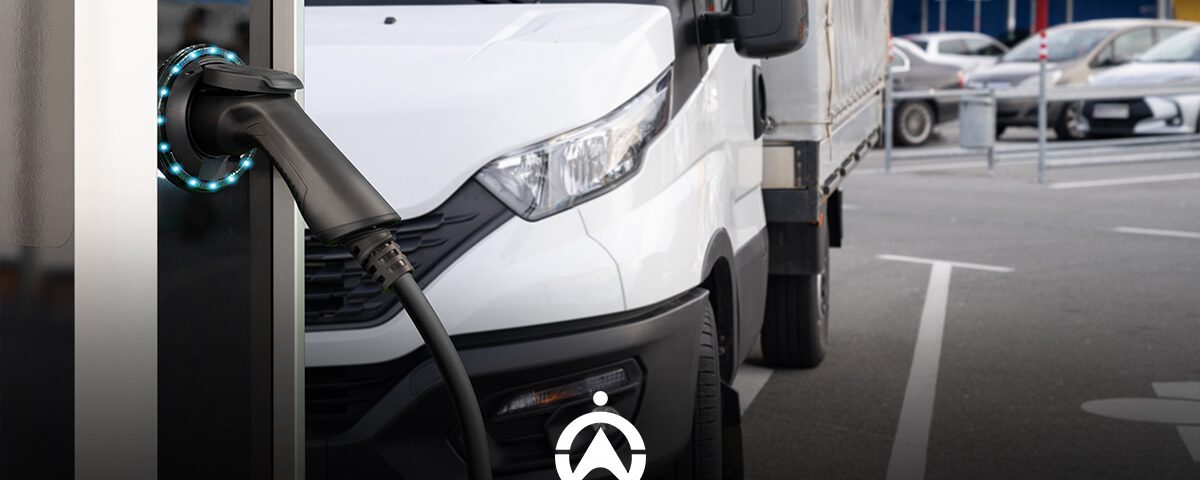 An electric vehicle charging station with a charging cable plugged in, with a parked white delivery truck in the background.