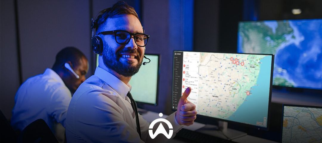 A person in a headset gives a thumbs up while operating multiple computer screens displaying maps and data in a control room.