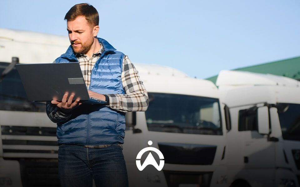 A person in a blue vest checks a laptop in front of white trucks against a clear sky, indicating a busy logistical environment.