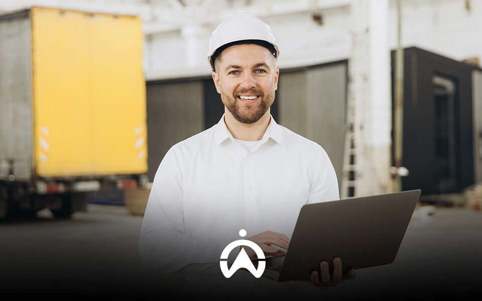 A construction worker in a white shirt and hard hat stands holding a laptop in a warehouse with shipping containers in the background.