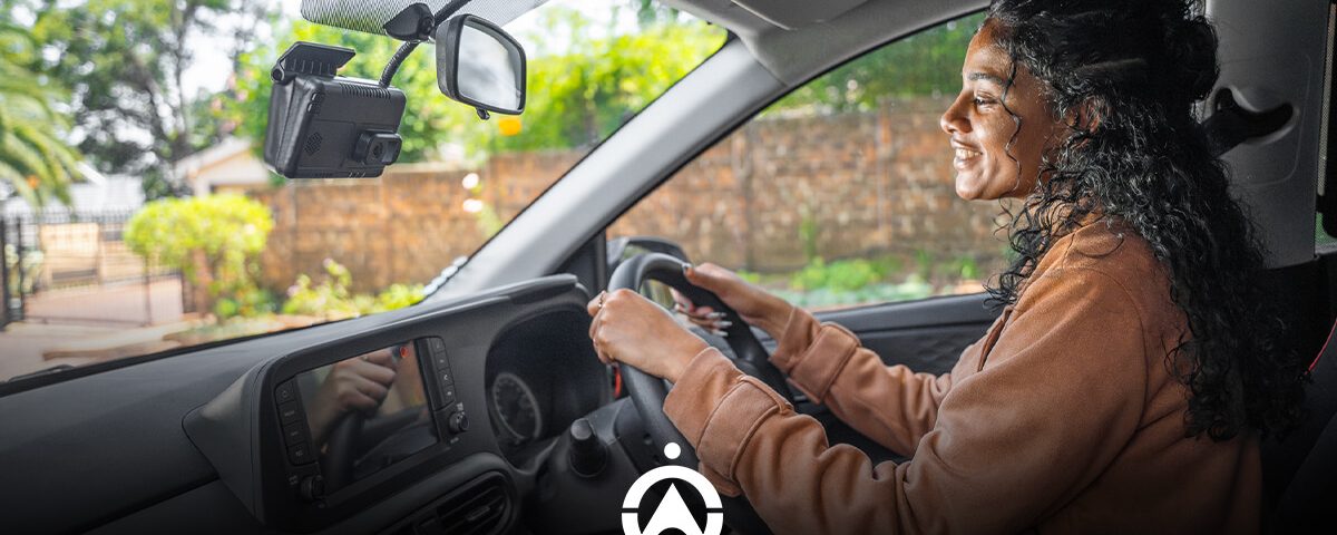 A person with curly hair drives a car, focused on the road ahead, with a dashboard camera mounted on the windshield.