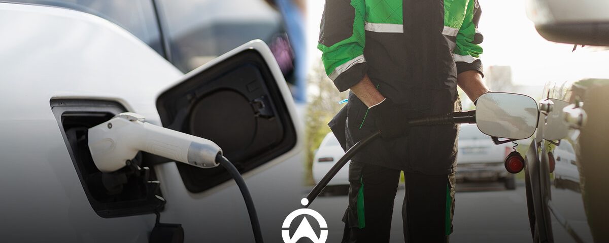 A technician wearing a black and green uniform refuels a vehicle while an electric charging cable is connected to another car.